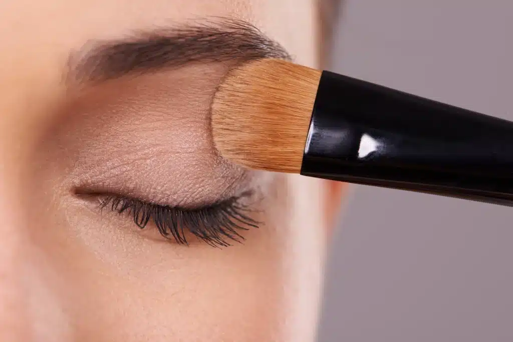 Maquillage des yeux : Appliquer son fard à paupières Pinceau appliquant de l'ombre à paupières beige sur l'œil fermé d'une femme pour un maquillage naturel.